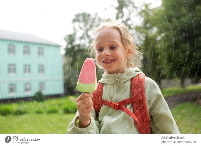 Positives Kind mit Rucksack, das im grünen Park steht und ein Eis bei Tageslicht genießt Speiseeis Kindheit Gras Glück essen niedlich bezaubernd genießen