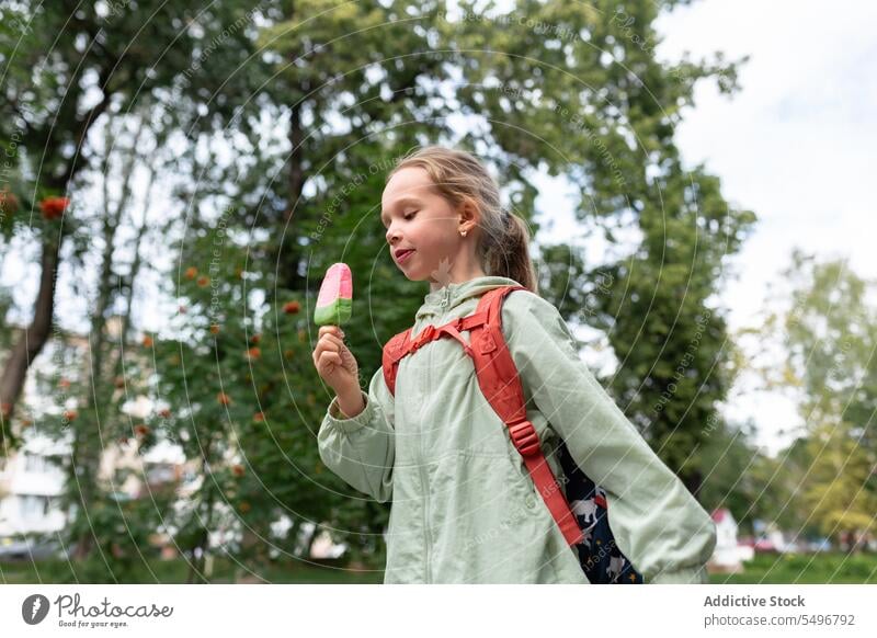 Glückliches Kind mit Rucksack im grünen Park stehend und ein Eis im Tageslicht genießend Speiseeis Kindheit Gras essen niedlich bezaubernd Mädchen Feld
