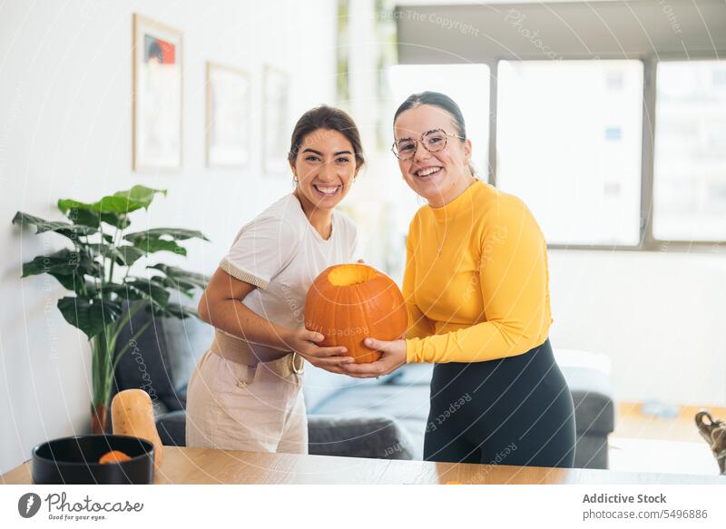 Glückliche ethnische junge Frauen stehen mit orangefarbenen Kürbis in den Händen zu Hause Freund Lächeln heiter positiv feiern Tisch hispanisch Brille Streifen