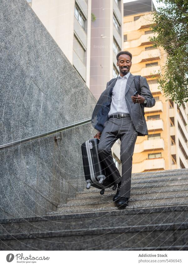 Fröhlicher schwarzer Mann mit Koffer geht die Treppe hinunter und surft auf seinem Smartphone im unteren Stockwerk unterirdisch Telefonanruf Glück formal