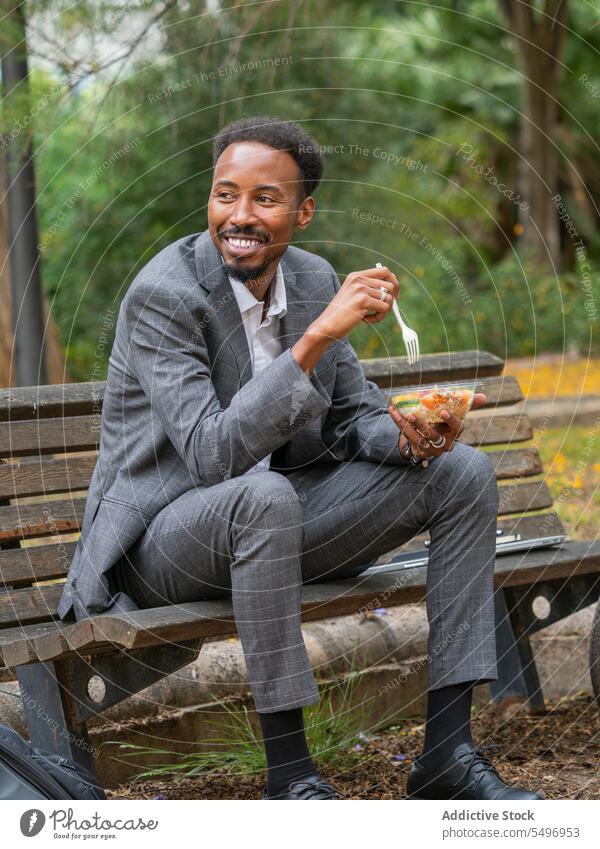 Glücklicher schwarzer Geschäftsmann sitzt auf einer Bank im Tageslicht und isst Salat in einer Schüssel Mann essen Salatbeilage Mittagessen Schalen & Schüsseln