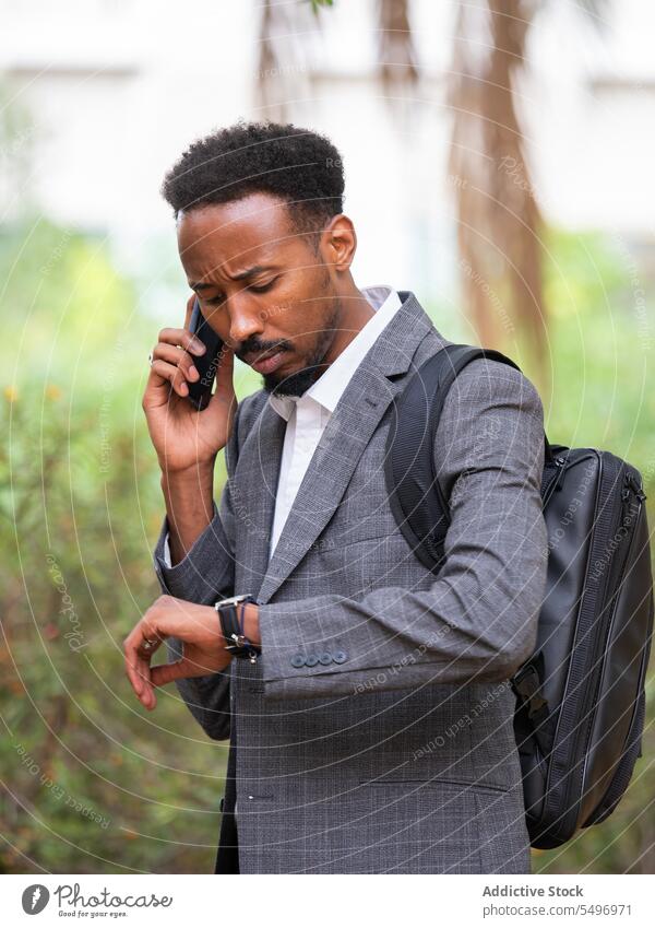 Stirnrunzelnder schwarzer Geschäftsmann, der mit seinem Smartphone spricht, während er bei Tageslicht die Uhrzeit überprüft reden selbstbewusst Telefonanruf