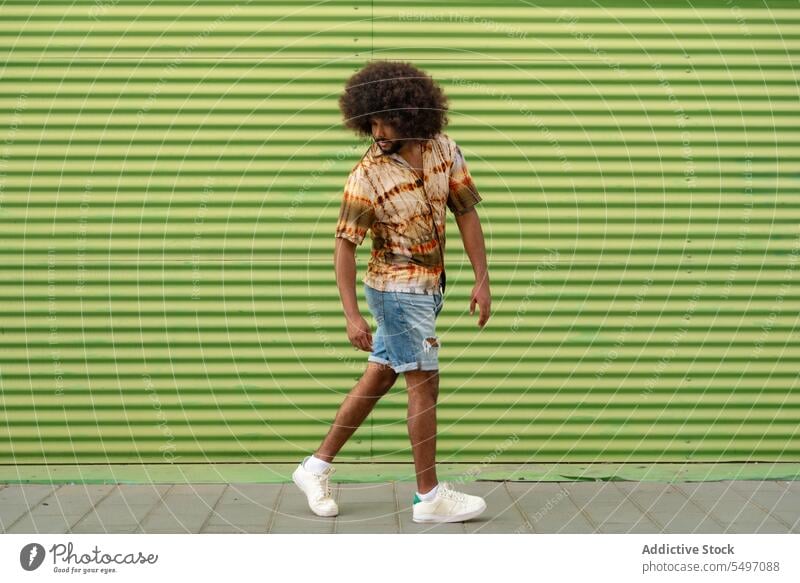 Junger ethnischer Mann geht auf dem Gehweg in der Nähe der Mauer Spaziergang Stil Straße ernst urban grün Streifen Wand trendy männlich lässig jung