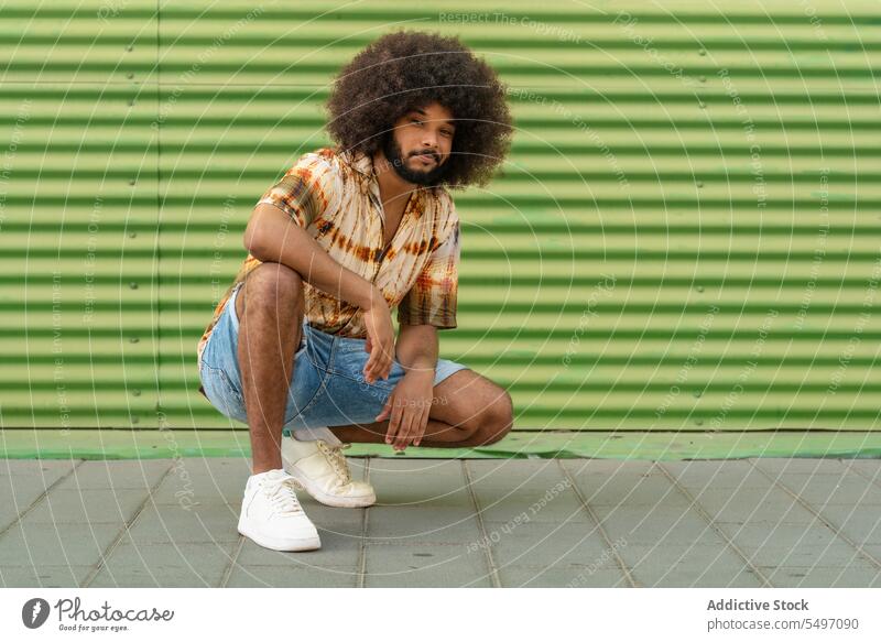 Stilvoller ethnischer Mann mit Afro-Haar vor gestreifter Wand trendy selbstbewusst Afro-Look cool krause Haare unabhängig Individualität hocken Outfit Vorschein