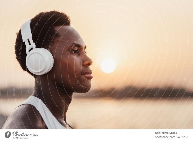 Schwarzer Mann hört Musik mit Kopfhörern in der Nähe von Wasser zuhören Sonnenuntergang ernst Drahtlos Gesang Gerät männlich Wiedergabeliste Apparatur