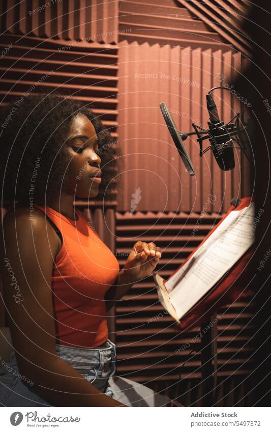Fokussierte schwarze Frau, die in der Nähe von Mikrofonen im Studio steht und ein Lied aufnimmt Sänger Atelier Aufzeichnen Gesang Papier Audio singen jung