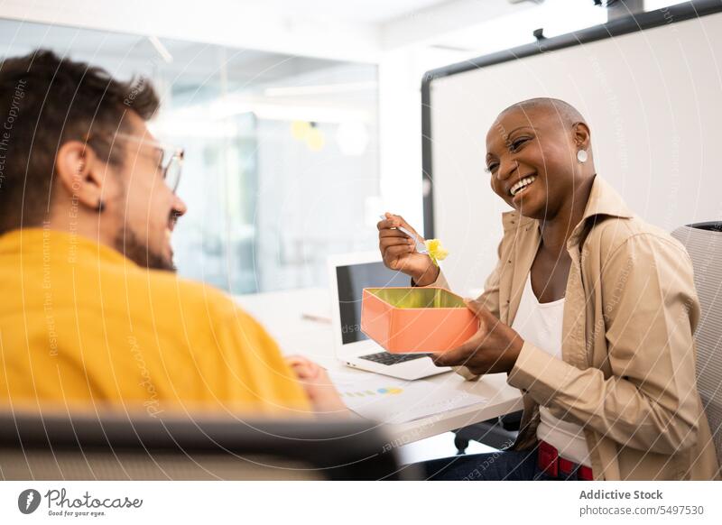 Fröhliche afroamerikanische Frau, die mit einem Freund an einem Tisch sitzt und im Büro zu Mittag isst Mann Kollege Lächeln Mittagessen Kasten Glück Pause