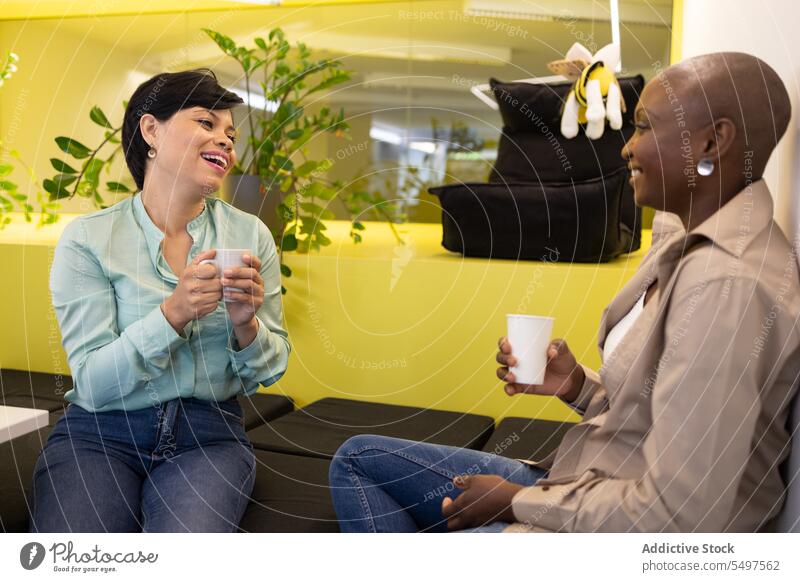 Fröhliche, gemischtrassige Kollegen, die sich auf Polsterstühlen in einem Raum mit Kaffeetassen ausruhen Frauen Kaffeepause reden Talkrunde Kommunizieren