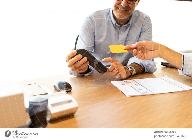 Lächelnder erwachsener Mann an einem Tisch mit einer POS-Maschine und einer unerkennbaren Frau, die eine Kreditkarte aushändigt Werkstatt Zahlung Kauf bezahlen