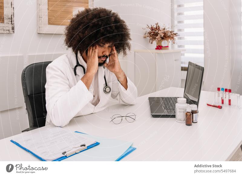 Nachdenklicher schwarzer Arzt sitzt am Schreibtisch im Krankenhaus Mann Stethoskop medizinisch Klinik besinnlich nachdenklich sich auf die Hand lehnen