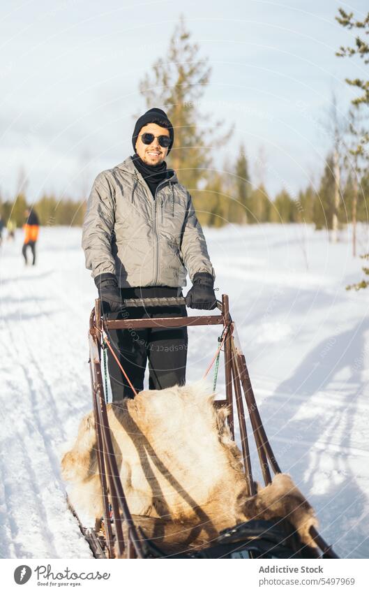 Fröhlicher Mann mit Sonnenbrille in der Nähe des Schlittens Reisender Rodel Winter Wald Schnee Straße froh Porträt männlich Tourist Lappland nordisch Urlaub
