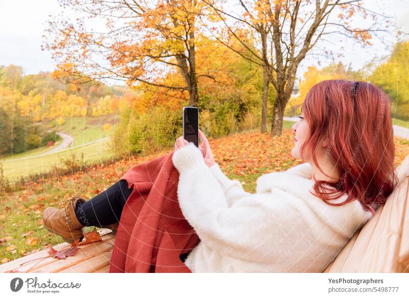 Frau benutzt ein Smartphone in einer Herbstlandschaft im Freien allein schön Schönheit Bank Decke Stiefel hell lässig Farbe farbenfroh genießen fallen Laubwerk