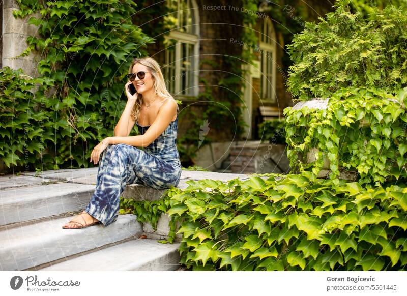 Hübsche junge Frau, die ein Handy benutzt, neben einem alten Haus mit Efeu Sonnenbrille Sitzen niedlich Kaukasier Erwachsener attraktiv schön Anruf Funktelefon