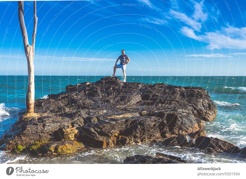 Mann mittleren Alters und Brille steht in heroischer Pose auf einem Fels im Meer Meeresufer Meeresküste felsig Felsen Wellen Wellengang Küste Küstenlandschaft