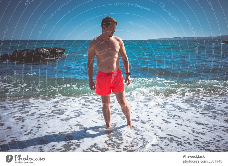 Mann mittleren Alters mit leuchtend roter Badehose steht im Meer und schaut zur Seite Sommer Sommerurlaub Sommertag Baden gehen Meeresufer Meerwasser Küste