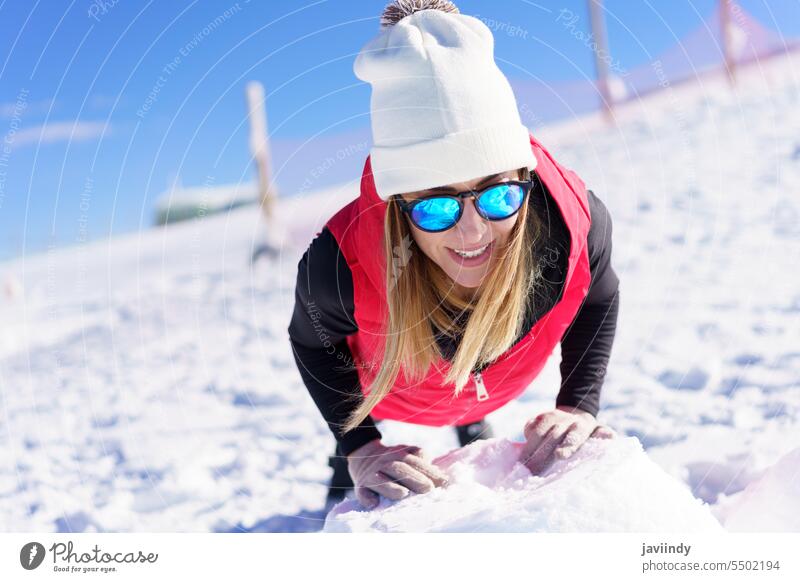 Lächelnde junge Frau mit Wollmütze und Sonnenbrille wärmt sich im Schnee auf anlehnen hochschieben Felsen passen Übung Glück positiv blond Verschlussdeckel