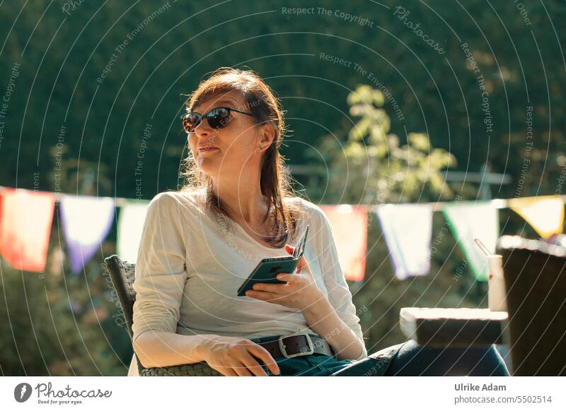 Drinkje bej Inkje |Sonnenstrahlen -  Frau mit Sonnenbrille relaxt und genießt im schönen Garten das sonnige Wetter. In der Hand hält sie ein Smartphone