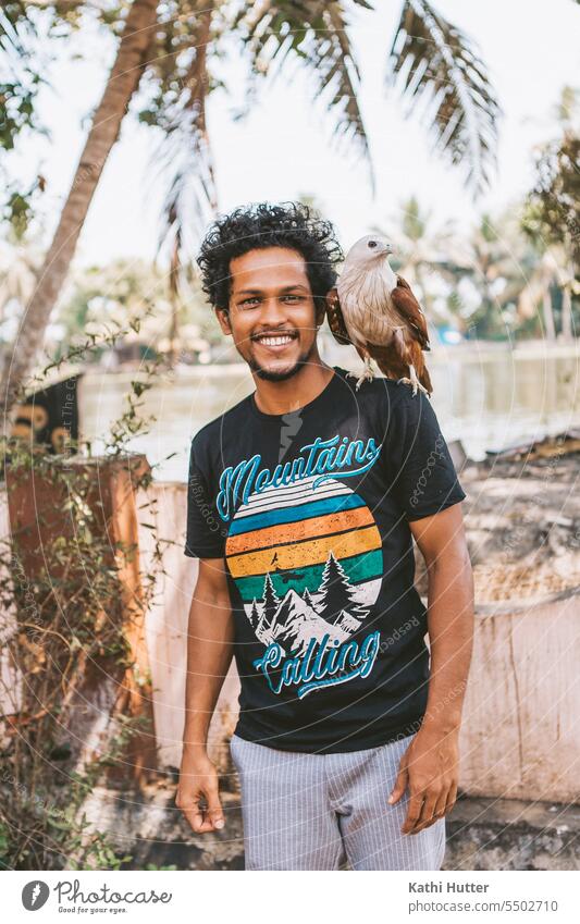 ein Adler sitzt auf der Schulter eines jungen Mannes, im Hintergrund sind einige Palmen zu sehen Männer junge Männer männlich Glück Lächeln Person Vogel