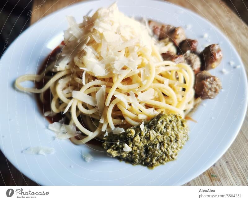 Spaghetti mit Bratwürstchen, Parmesanflocken und grünem Pesto auf einem weißen Teller, der auf einem hölzernen Schneidebrett steht Pasta Nudeln Teigwaren