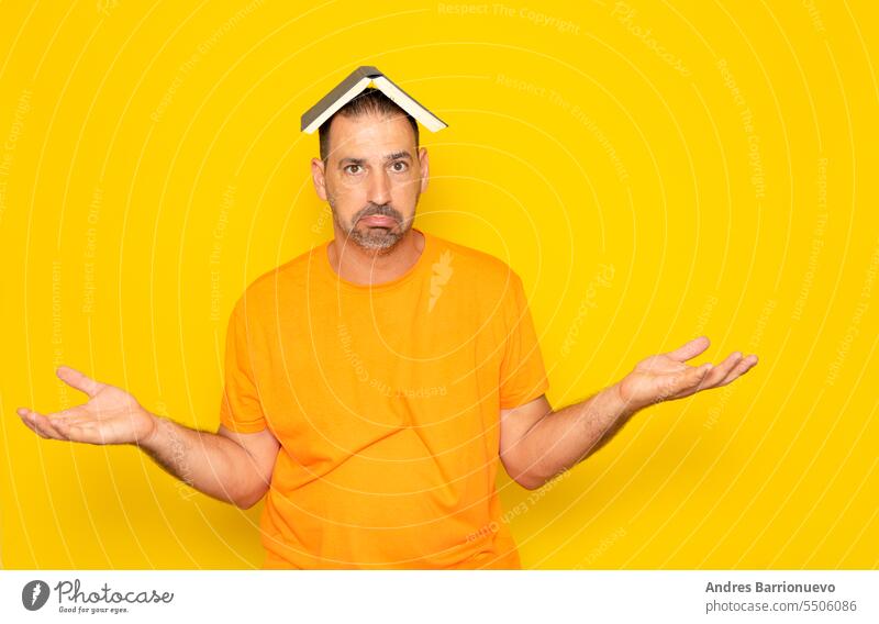 Bärtiger hispanischer Mann in den 40ern, der ein orangefarbenes T-Shirt trägt und ein aufgeschlagenes Buch auf dem Kopf hält und die Hände hebt, als würde er nichts verstehen, isoliert auf gelbem Studiohintergrund