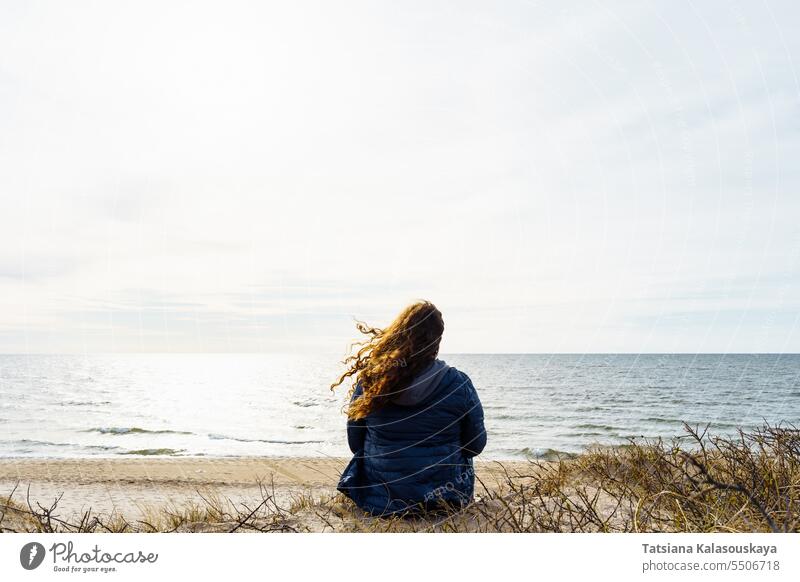 Lockenköpfige Frau in dünner Daunenjacke sitzt abends am Meer, die Haare flattern im Wind Lockenhaarige Übergröße dünne Daunenjacke Meeresufer Ostsee Abend