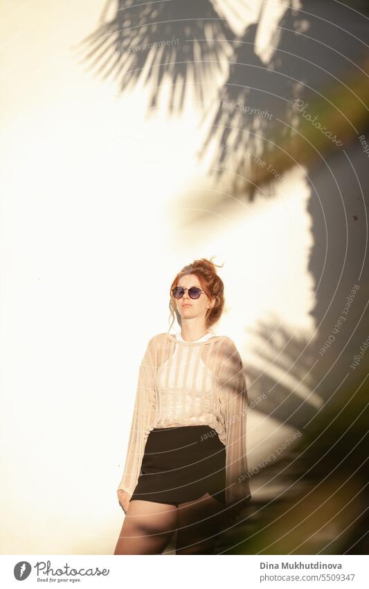 junge schöne rothaarige Frau in Freizeitkleidung mit Sonnenbrille und Haarknoten auf beigem Wandhintergrund mit Palmenschatten. Candid Outdoor-Porträt im Park.