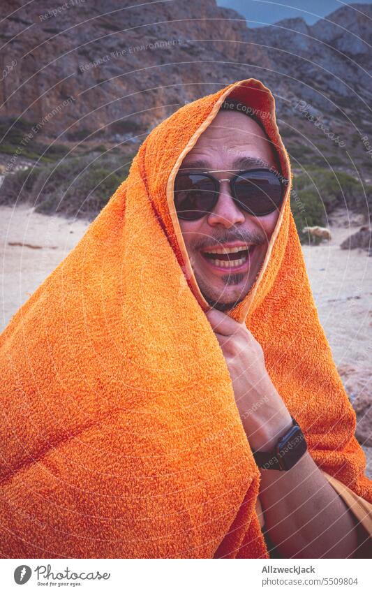 Mann mittleren Alters mit Sonnenbrille sitzt in ein Handtuch gewickelt am Strand und lacht Sommer Sommerurlaub Urlaub Urlaubsstimmung Urlaubsfoto sommerlich