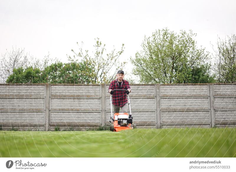 Blick auf Mann in Freizeitkleidung mäht Rasen mit Rasenmäher im Hinterhof seines Hauses. Der Ehemann kümmert sich um den Garten an einem bewölkten Frühlingstag. Moderne Benzin-Gartengeräte. Landschaftsbau Arbeit