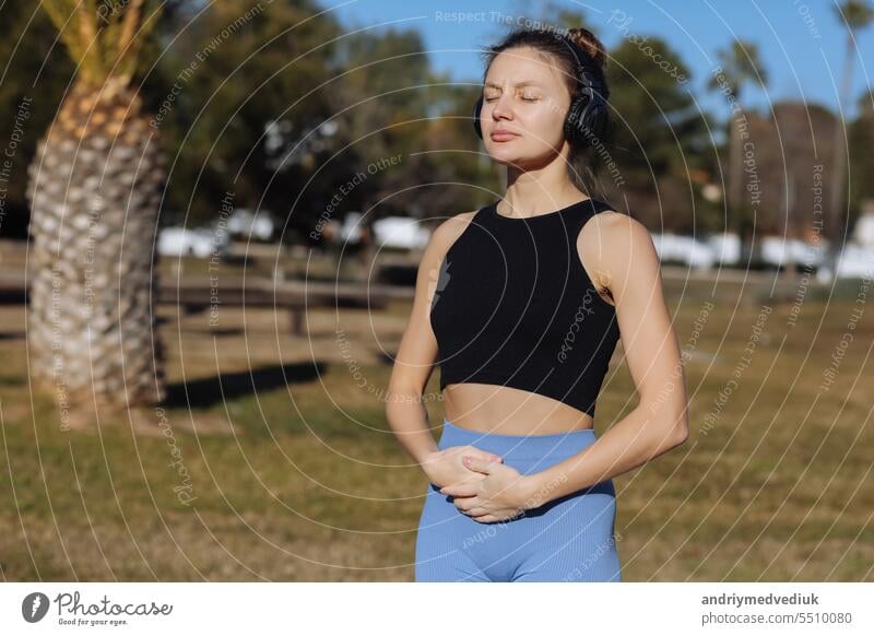 Konzentrierte junge Frau mit Kopfhörern macht Yoga-Meditation mit Online-Tutorial im Freien im Park mit Palmen. Hören von Musik, Harmonie, Beruhigung, Spiritualität Konzept. Weltgesundheitstag