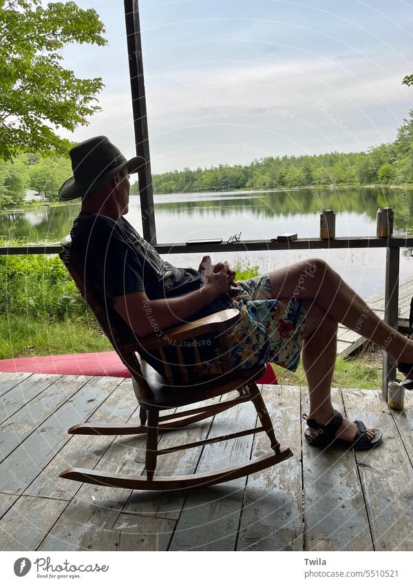 Mann entspannt sich im Schaukelstuhl auf der Terrasse einer Hütte vor einem See Veranda im Freien Außenaufnahme Erholung Sommer Windstille