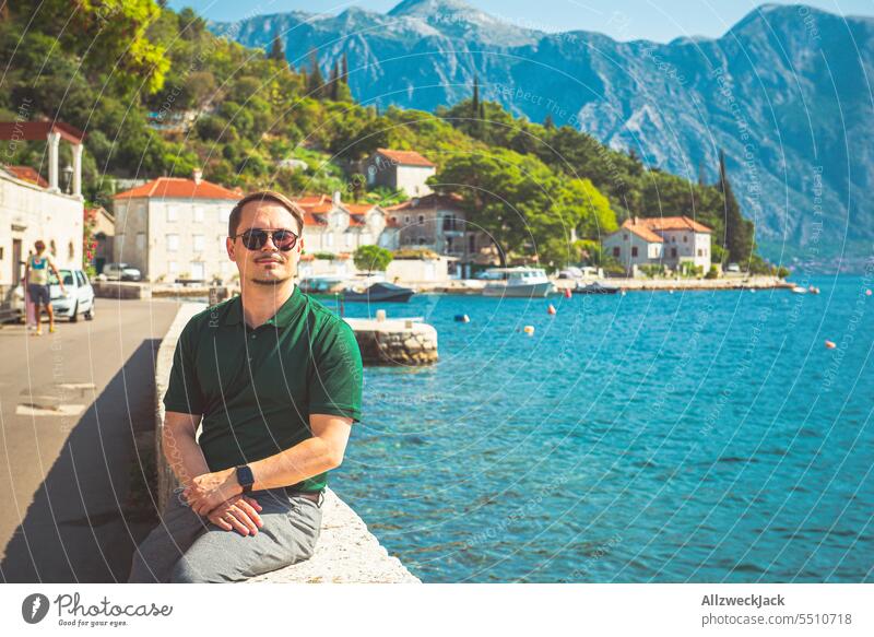 Mann mittleren Alters mit grünem Shirt und Sonnenbrille sitzt auf einer Mauer an der Küste von Perast in Montenegro Mittelmeer mittelmeerraum Wasser