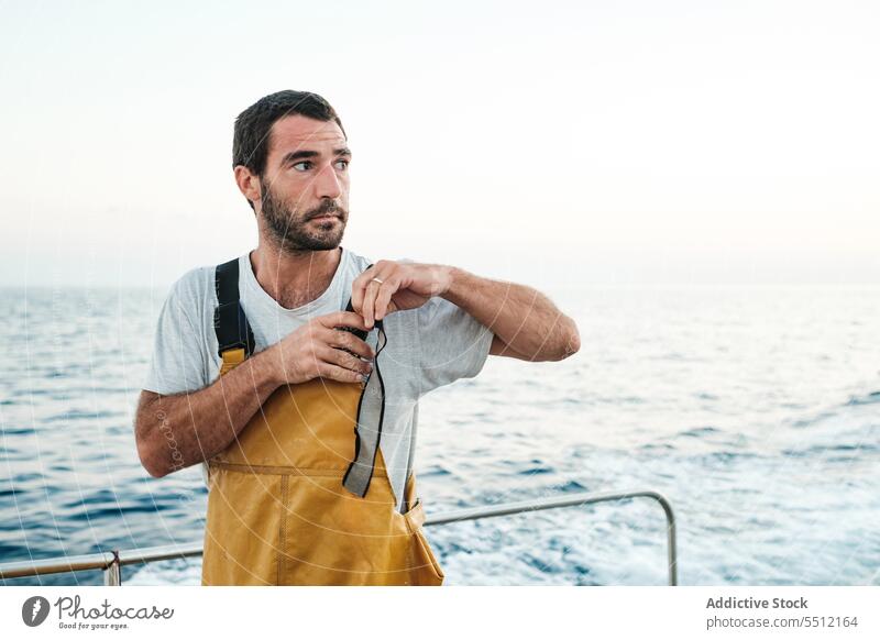 Selbstbewusster junger ethnischer Fischer, der die Hosenträger seines Lätzchens auf dem Segelboot festzieht Mann Boot reisen befestigen wasserdicht Klammer