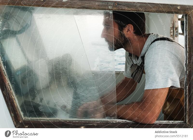 Bärtiger ethnischer Mann in Latzhose sitzt in einer Bootskabine in der Nähe des Armaturenbretts Kabine MEER Fenster reisen Segel Wasser Verkehr Sommer männlich