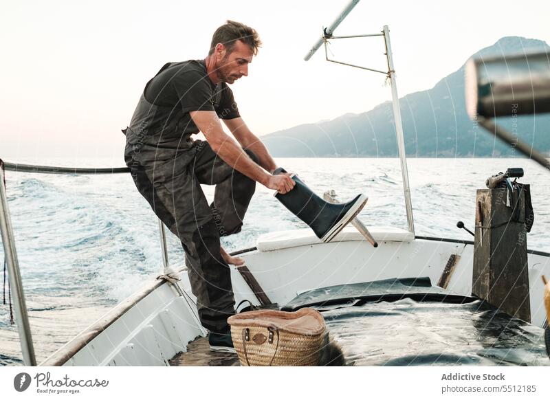 Junger ethnischer Mann steht auf einem Boot und trägt Gummisegelschuhe Segelboot MEER Abend Reling Meereslandschaft Zaun Metall anlehnen Meerwasser männlich