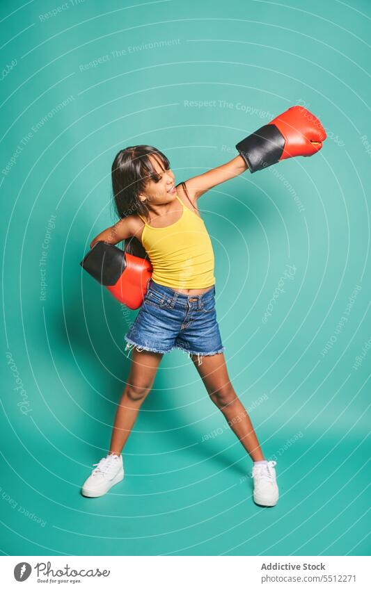 Fröhliches ethnisches Mädchen in Boxhandschuhen, das Schläge vor türkisfarbenem Hintergrund austeilt Kind Lächeln Kindheit Spaß haben Boxsport Handschuh Glück