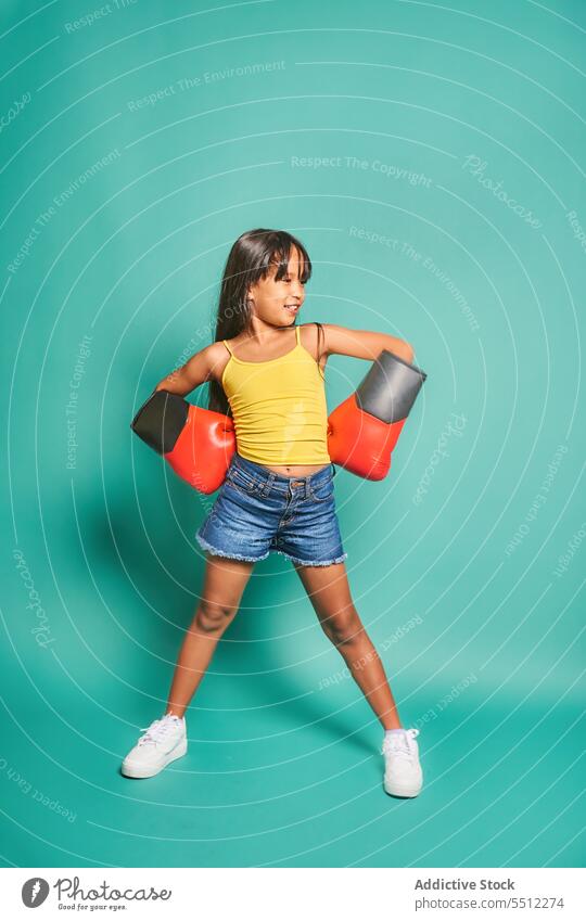 Fröhliches ethnisches Mädchen mit Boxhandschuhen vor türkisfarbenem Hintergrund Kind Lächeln Kindheit Spaß haben Boxsport Handschuh Glück werfen spielerisch