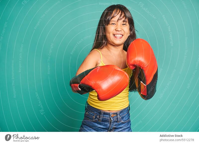 Fröhliches ethnisches Mädchen mit Boxhandschuhen vor türkisfarbenem Hintergrund Kind Lächeln Boxsport Handschuh Porträt froh positiv heiter Raum spielerisch