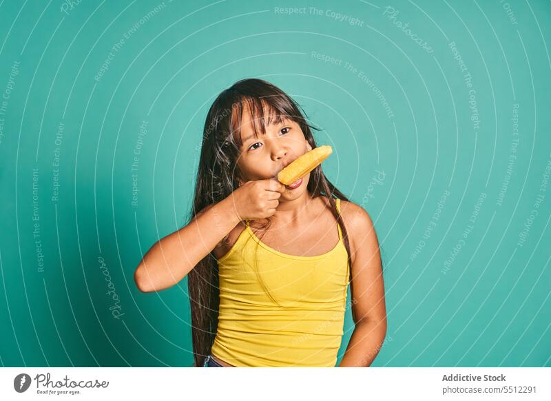 Glückliches ethnisches Mädchen Kind steht und isst Eis in der Hand gegen türkisfarbenen Hintergrund Stieleis lecker Vorschein süß niedlich geschmackvoll
