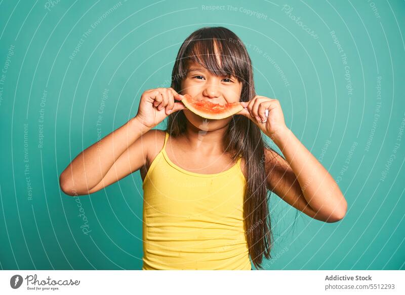 Glückliches ethnisches Mädchen Kind steht und isst ein Stück Wassermelone vor türkisem Hintergrund lecker Vorschein süß niedlich geschmackvoll bezaubernd