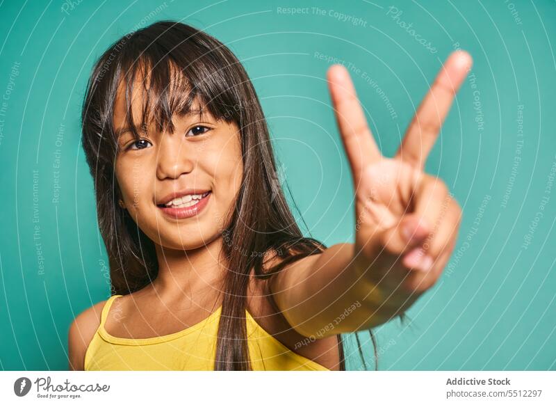 Glückliches ethnisches Mädchen, das vor einem türkisfarbenen Hintergrund steht und das Siegeszeichen zeigt gestikulieren Lächeln Kind v-Zeichen zwei Finger
