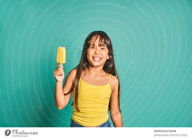Glückliches ethnisches Mädchen mit einem Eis in der Hand vor einem türkisfarbenen Hintergrund Kind Stieleis Kindheit zeigen gefroren niedlich lecker spielerisch