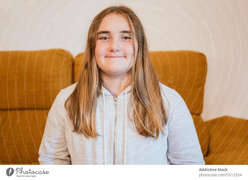 Lächelndes junges Mädchen im Kapuzenpulli sitzt auf einem Stuhl im Wohnzimmer positiv Sofa Porträt Vorschein Glück froh angenehm zu Hause Teenager Stil charmant
