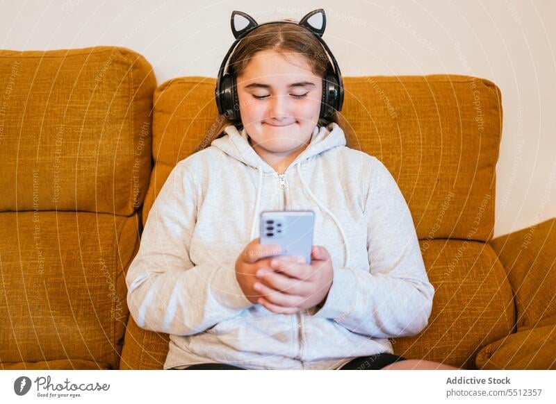 Lächelndes junges Mädchen mit Kopfhörern und Smartphone beim Musikhören zuhören benutzend Sofa online Glück Teenager Apparatur Gerät Freizeit Mobile Telefon