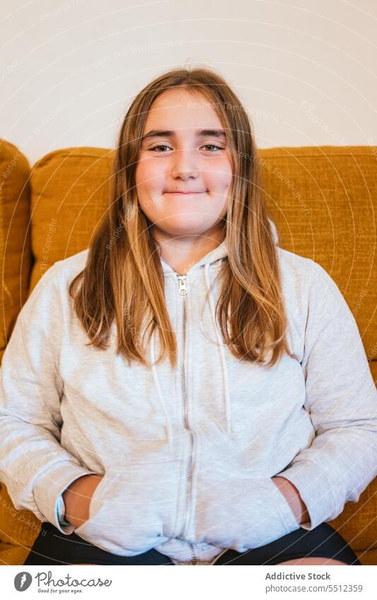 Lächelndes junges Mädchen im Kapuzenpulli sitzt auf einem Stuhl im Wohnzimmer positiv Sofa Porträt Vorschein Glück froh angenehm zu Hause Teenager heiter Stil