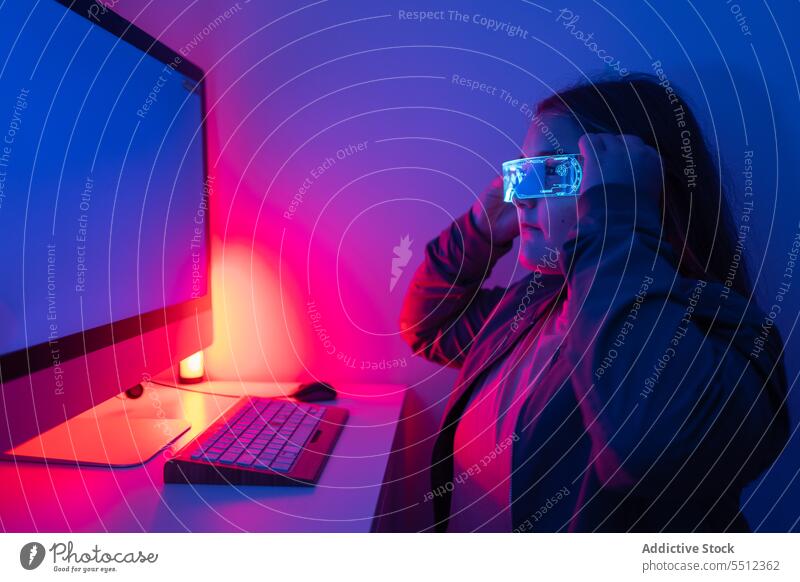 Junges Mädchen mit AR-Brille sitzt am Schreibtisch mit Computermonitor Teenager Bildschirm Monitor neonfarbig Tisch Eckstoß Raum Apparatur Gerät Licht Anzeige