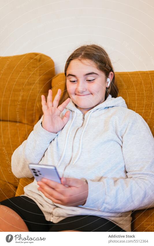 Fröhliches junges Mädchen, das auf dem Sofa sitzend sein Smartphone begrüßt Videoanruf Lächeln Hallo online benutzend Kommunizieren Anschluss Internet Teenager