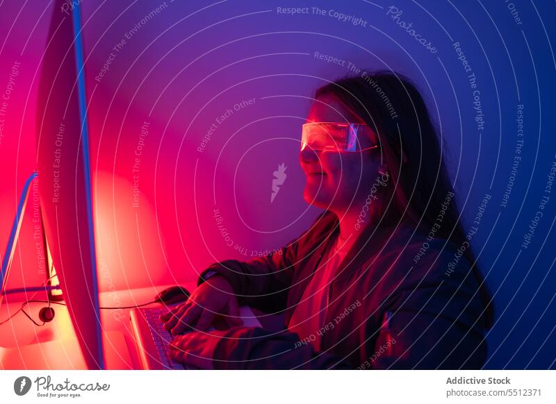Junges Mädchen mit AR-Brille sitzt am Schreibtisch mit Computermonitor Teenager Bildschirm Monitor neonfarbig Tisch Lächeln Eckstoß Raum Apparatur Gerät Licht