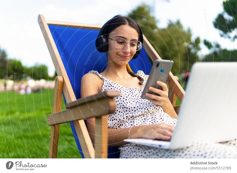 Lächelnde Frau mit Laptop und Smartphone auf einem Liegestuhl benutzend Park positiv Mobile Sommer Telefon Browsen jung Internet Surfen Glück Gerät Apparatur