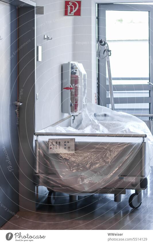 Ein ungenutztes, mit einer Folie bedecktes Krankenbett auf dem zugigen Gang einer Klinik Krankenhaus Gesundheit krank Krankheit Liege Bett Station Patient