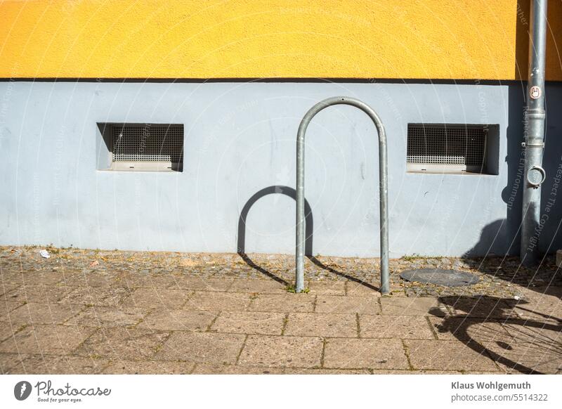 Licht und Schatten an einer Hauswand. Ein Fahrradständer aus verzinktem Stahlrohr und eine ebensolche Regenrinne stehen vor der gelb/blauen Fassade. Der Schatten eines Fahrrades schiebt sich von rechts ins Bild.